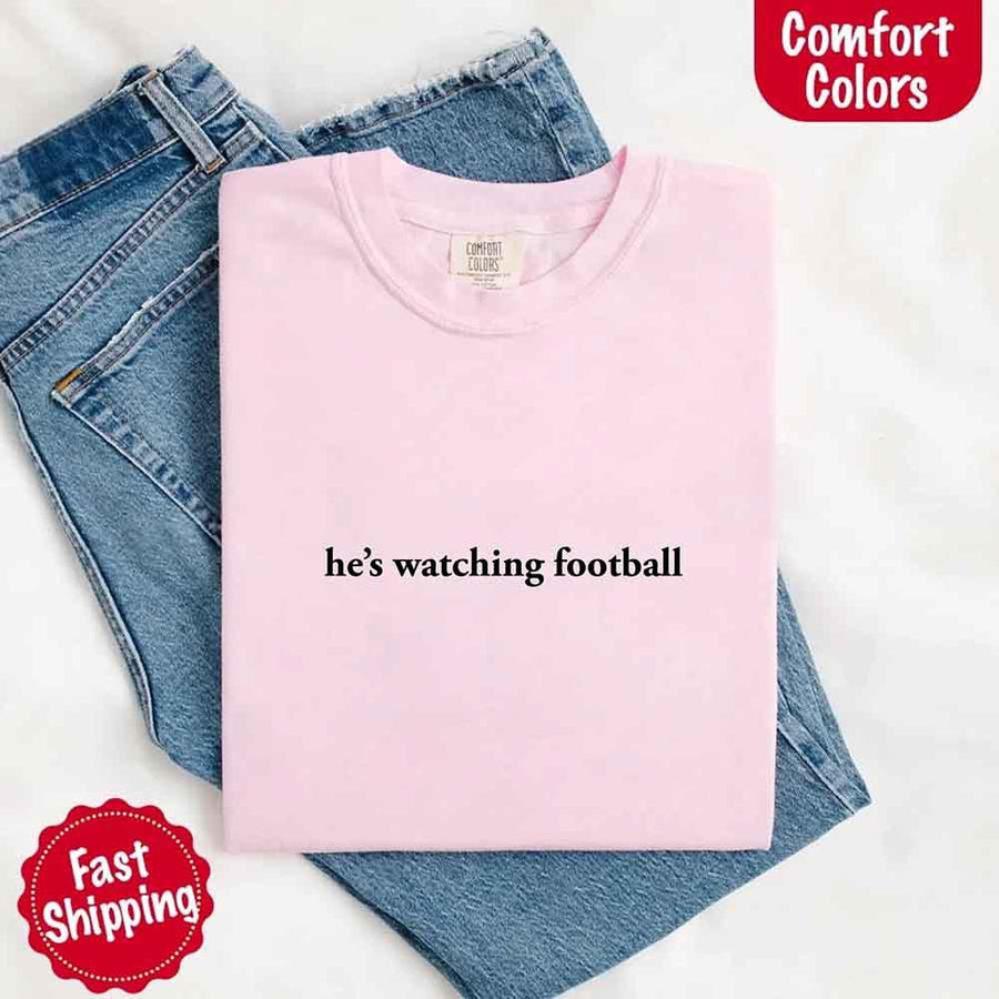 May include a woman wearing an ivory Comfort Colors t-shirt with the phrase 'He’s Watching Football' — styled in a comfy, casual look perfect for Sundays or football season humor