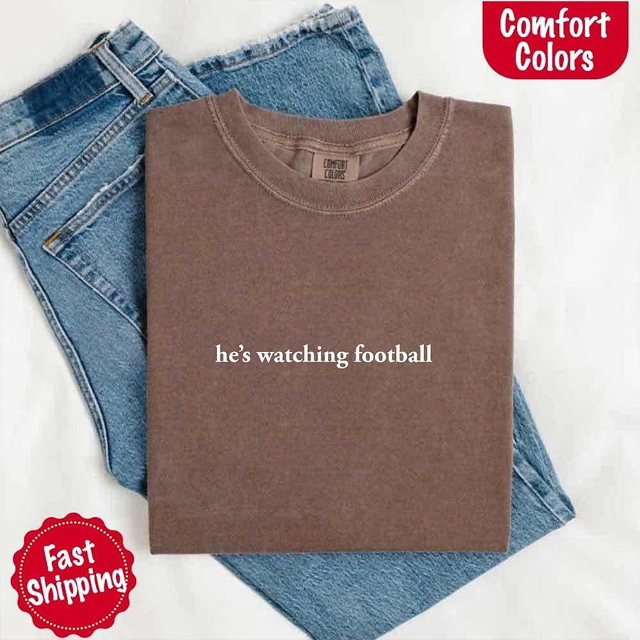 May include a woman wearing an ivory Comfort Colors t-shirt with the phrase 'He’s Watching Football' — styled in a comfy, casual look perfect for Sundays or football season humor