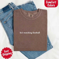 May include a woman wearing an ivory Comfort Colors t-shirt with the phrase 'He’s Watching Football' — styled in a comfy, casual look perfect for Sundays or football season humor