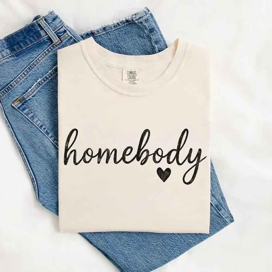 May include a folded ivory Comfort Colors “homebody” t-shirt in neutral tones, with minimalist black lettering and heart detail, laid on denim jeans.
