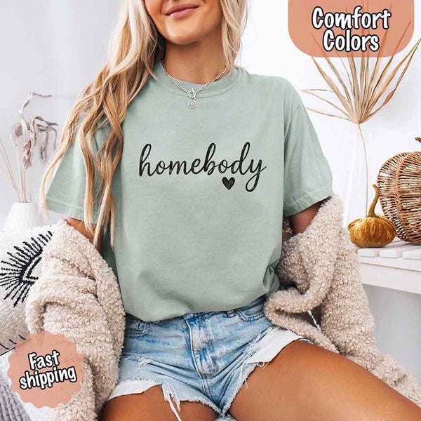 May include a woman wearing a bay Comfort Colors “homebody” t-shirt with black script text and heart graphic, styled casually with denim shorts and cozy cardigan.