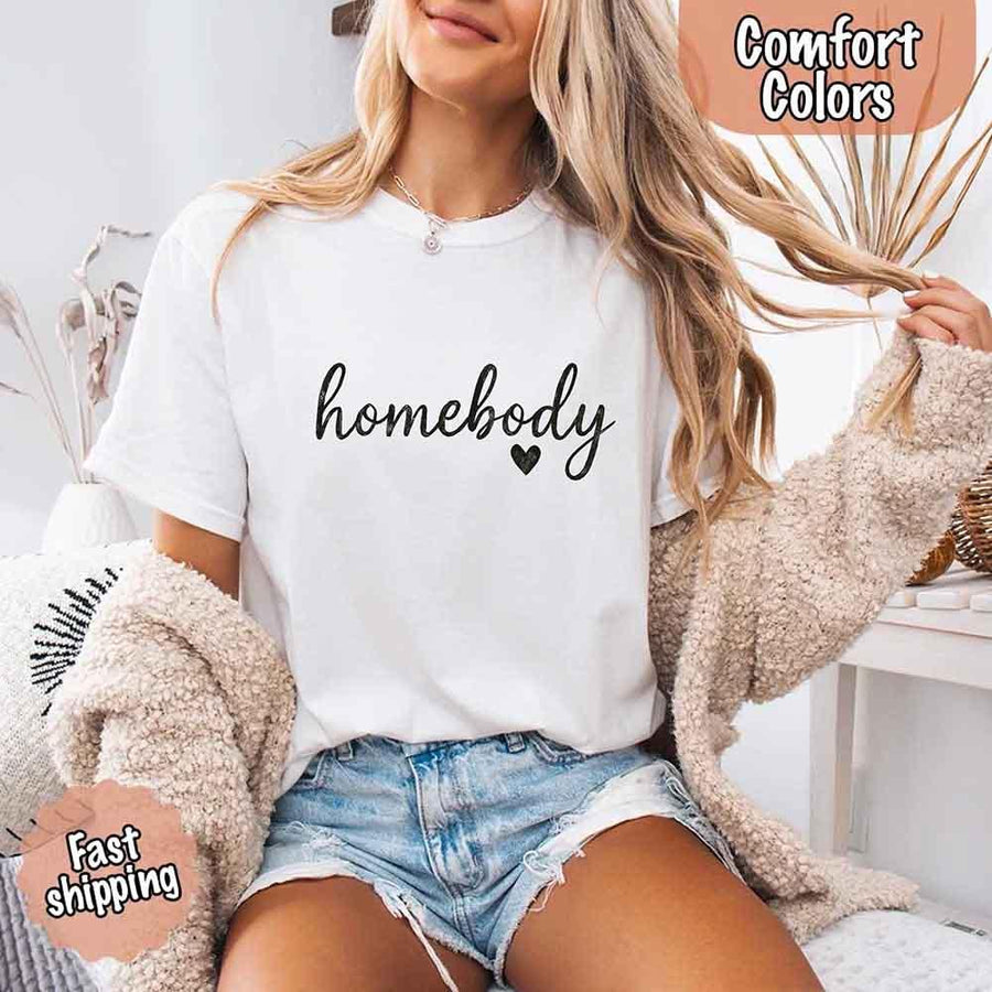 May include a woman wearing a  white Comfort Colors “homebody” t-shirt with black script text and heart graphic, styled casually with denim shorts and cozy cardigan.