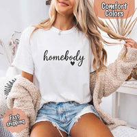 May include a woman wearing a  white Comfort Colors “homebody” t-shirt with black script text and heart graphic, styled casually with denim shorts and cozy cardigan.