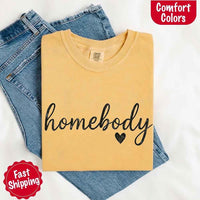 May include a folded mustard Comfort Colors “homebody” t-shirt in neutral tones, with minimalist black lettering and heart detail, laid on denim jeans.