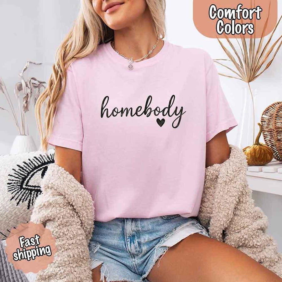 May include a woman wearing a blossom Comfort Colors “homebody” t-shirt with black script text and heart graphic, styled casually with denim shorts and cozy cardigan.