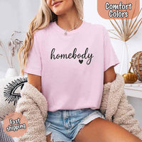 May include a woman wearing a blossom Comfort Colors “homebody” t-shirt with black script text and heart graphic, styled casually with denim shorts and cozy cardigan.