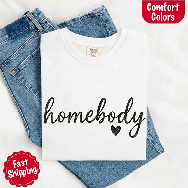 May include a folded white Comfort Colors “homebody” t-shirt in neutral tones, with minimalist black lettering and heart detail, laid on denim jeans.