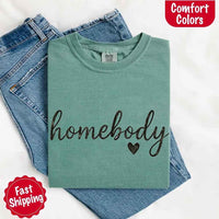 May include a folded light green Comfort Colors “homebody” t-shirt in neutral tones, with minimalist black lettering and heart detail, laid on denim jeans.