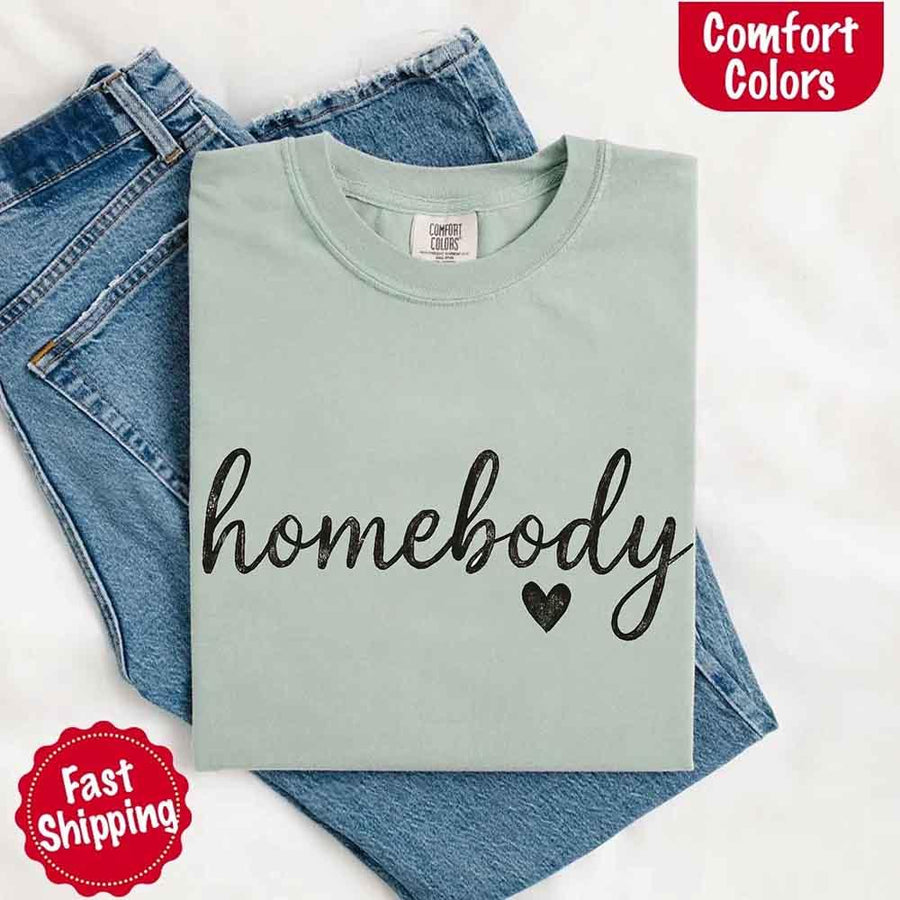 May include a folded bay Comfort Colors “homebody” t-shirt in neutral tones, with minimalist black lettering and heart detail, laid on denim jeans.
