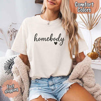 May include a woman wearing an ivory Comfort Colors “homebody” t-shirt with black script text and heart graphic, styled casually with denim shorts and cozy cardigan.