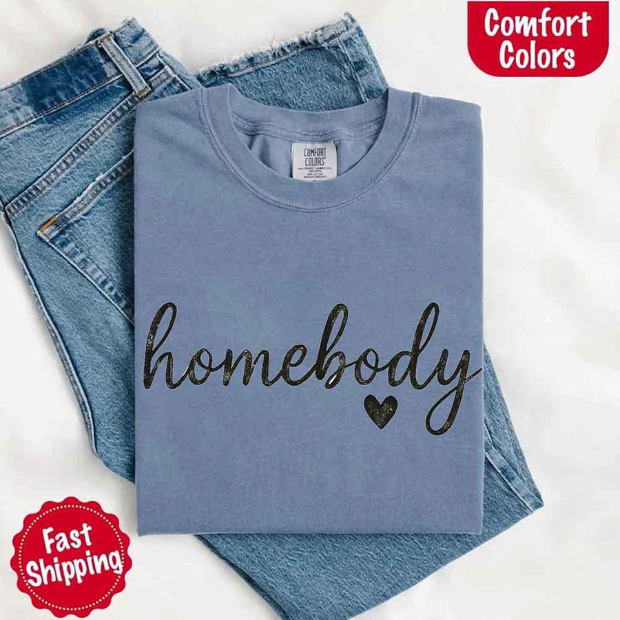 May include a folded blue jean Comfort Colors “homebody” t-shirt in neutral tones, with minimalist black lettering and heart detail, laid on denim jeans.