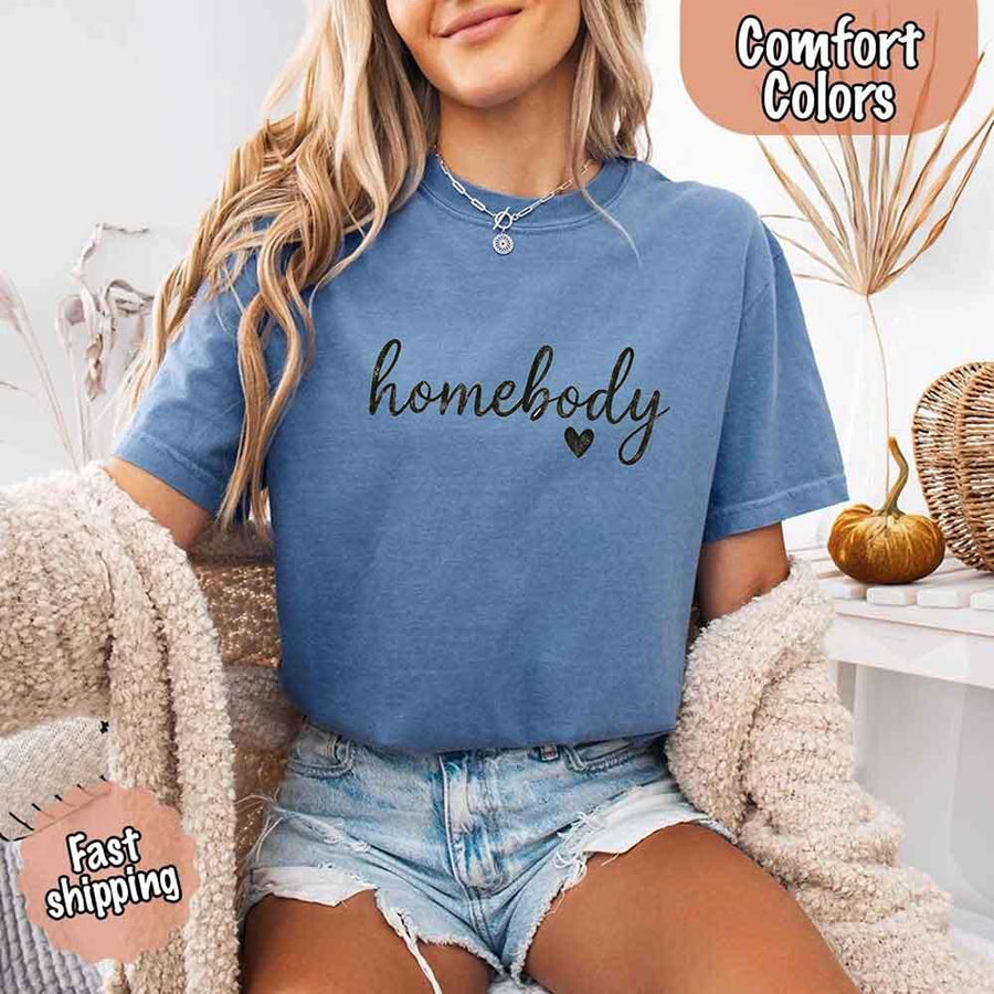 May include a woman wearing a blue jean Comfort Colors “homebody” t-shirt with black script text and heart graphic, styled casually with denim shorts and cozy cardigan.