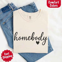 May include a folded ivory Comfort Colors “homebody” t-shirt in neutral tones, with minimalist black lettering and heart detail, laid on denim jeans.