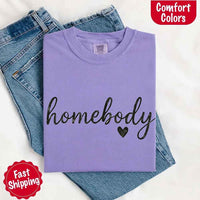 May include a folded violet Comfort Colors “homebody” t-shirt in neutral tones, with minimalist black lettering and heart detail, laid on denim jeans.