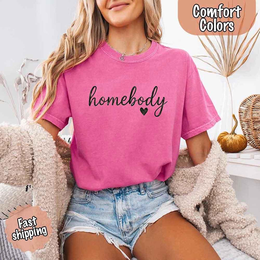May include a woman wearing a crunchberry Comfort Colors “homebody” t-shirt with black script text and heart graphic, styled casually with denim shorts and cozy cardigan.