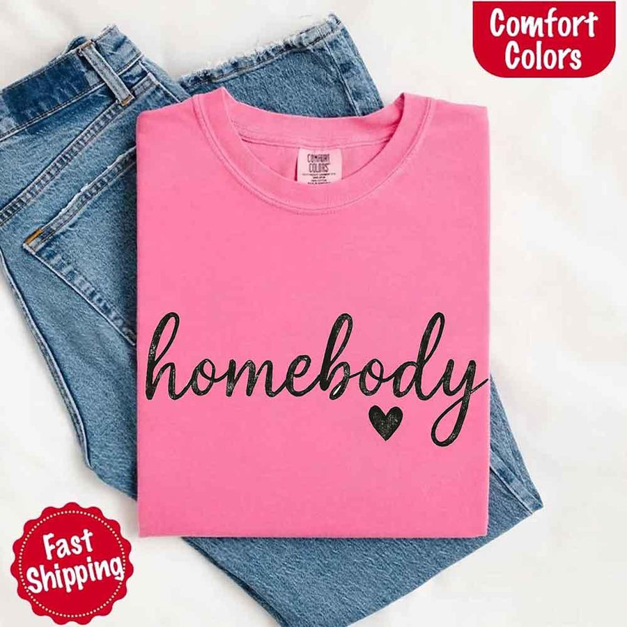 May include a folded crunchberry Comfort Colors “homebody” t-shirt in neutral tones, with minimalist black lettering and heart detail, laid on denim jeans.