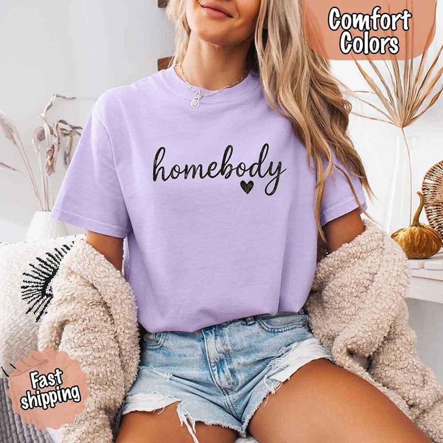 May include a woman wearing an orchid Comfort Colors “homebody” t-shirt with black script text and heart graphic, styled casually with denim shorts and cozy cardigan. 