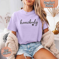 May include a woman wearing an orchid Comfort Colors “homebody” t-shirt with black script text and heart graphic, styled casually with denim shorts and cozy cardigan. 