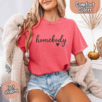 May include a woman wearing a watermelon Comfort Colors “homebody” t-shirt with black script text and heart graphic, styled casually with denim shorts and cozy cardigan.