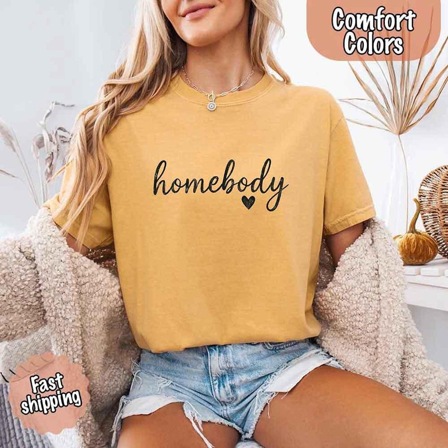 May include a woman wearing a mustard Comfort Colors “homebody” t-shirt with black script text and heart graphic, styled casually with denim shorts and cozy cardigan.