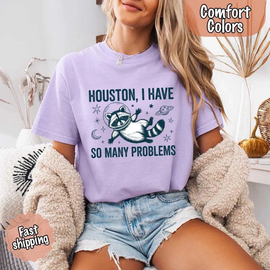 May include a cartoon raccoon in a space helmet floating in space, surrounded by stars, a planet with rings, and a crescent moon. The text reads: “HOUSTON, I HAVE SO MANY PROBLEMS,” parodying the famous Apollo 13 quote with a humorous twist.
