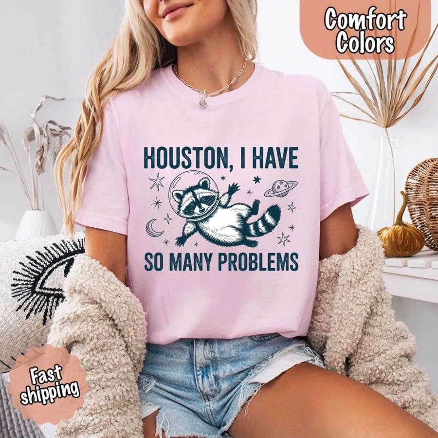 May include a cartoon raccoon in a space helmet floating in space, surrounded by stars, a planet with rings, and a crescent moon. The text reads: “HOUSTON, I HAVE SO MANY PROBLEMS,” parodying the famous Apollo 13 quote with a humorous twist.