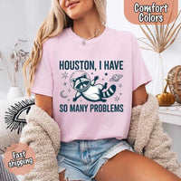 May include a cartoon raccoon in a space helmet floating in space, surrounded by stars, a planet with rings, and a crescent moon. The text reads: “HOUSTON, I HAVE SO MANY PROBLEMS,” parodying the famous Apollo 13 quote with a humorous twist.