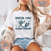 May include a cartoon raccoon in a space helmet floating in space, surrounded by stars, a planet with rings, and a crescent moon. The text reads: “HOUSTON, I HAVE SO MANY PROBLEMS,” parodying the famous Apollo 13 quote with a humorous twist.