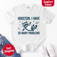 May include a cartoon raccoon in a space helmet floating in space, surrounded by stars, a planet with rings, and a crescent moon. The text reads: “HOUSTON, I HAVE SO MANY PROBLEMS,” parodying the famous Apollo 13 quote with a humorous twist.