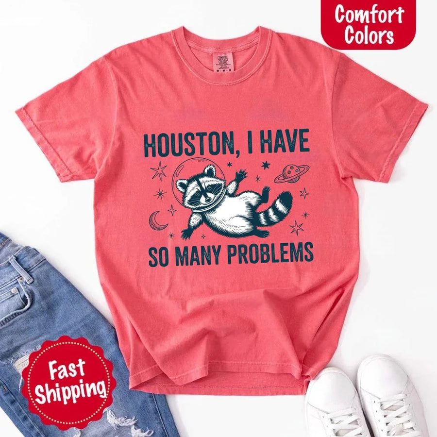 May include a cartoon raccoon in a space helmet floating in space, surrounded by stars, a planet with rings, and a crescent moon. The text reads: “HOUSTON, I HAVE SO MANY PROBLEMS,” parodying the famous Apollo 13 quote with a humorous twist.
