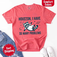 May include a cartoon raccoon in a space helmet floating in space, surrounded by stars, a planet with rings, and a crescent moon. The text reads: “HOUSTON, I HAVE SO MANY PROBLEMS,” parodying the famous Apollo 13 quote with a humorous twist.