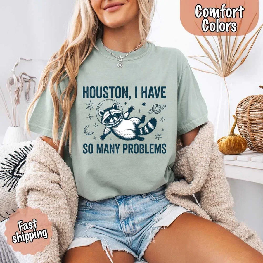 May include a cartoon raccoon in a space helmet floating in space, surrounded by stars, a planet with rings, and a crescent moon. The text reads: “HOUSTON, I HAVE SO MANY PROBLEMS,” parodying the famous Apollo 13 quote with a humorous twist.
