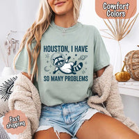 May include a cartoon raccoon in a space helmet floating in space, surrounded by stars, a planet with rings, and a crescent moon. The text reads: “HOUSTON, I HAVE SO MANY PROBLEMS,” parodying the famous Apollo 13 quote with a humorous twist.
