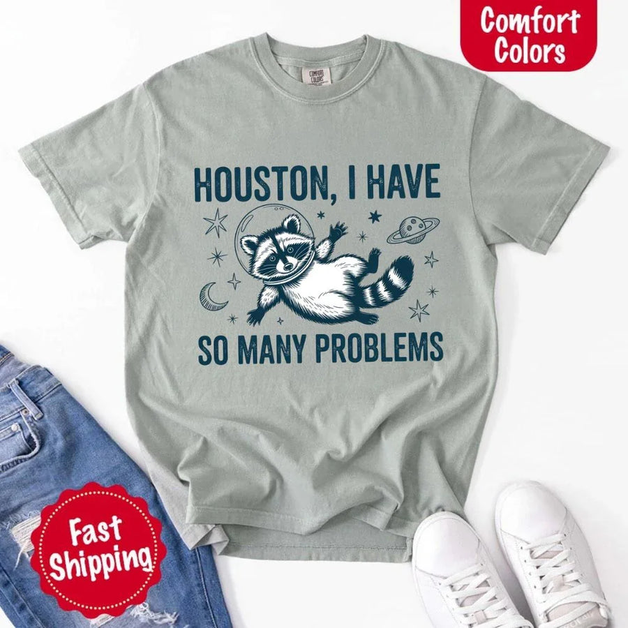 May include a cartoon raccoon in a space helmet floating in space, surrounded by stars, a planet with rings, and a crescent moon. The text reads: “HOUSTON, I HAVE SO MANY PROBLEMS,” parodying the famous Apollo 13 quote with a humorous twist.