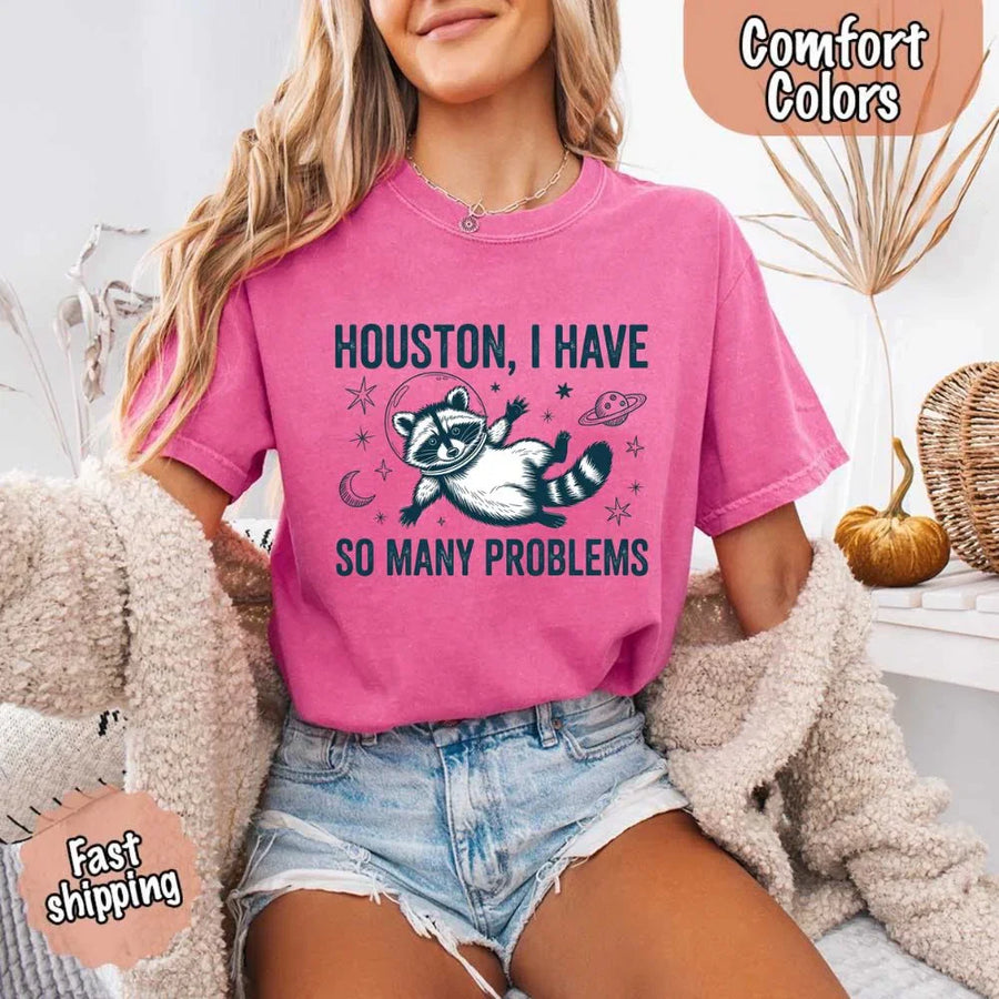 May include a cartoon raccoon in a space helmet floating in space, surrounded by stars, a planet with rings, and a crescent moon. The text reads: “HOUSTON, I HAVE SO MANY PROBLEMS,” parodying the famous Apollo 13 quote with a humorous twist.