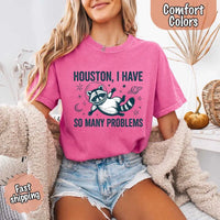 May include a cartoon raccoon in a space helmet floating in space, surrounded by stars, a planet with rings, and a crescent moon. The text reads: “HOUSTON, I HAVE SO MANY PROBLEMS,” parodying the famous Apollo 13 quote with a humorous twist.