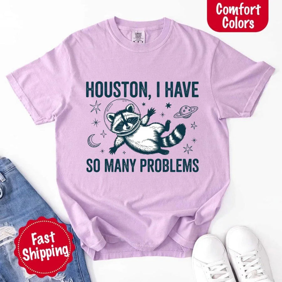 May include a cartoon raccoon in a space helmet floating in space, surrounded by stars, a planet with rings, and a crescent moon. The text reads: “HOUSTON, I HAVE SO MANY PROBLEMS,” parodying the famous Apollo 13 quote with a humorous twist.
