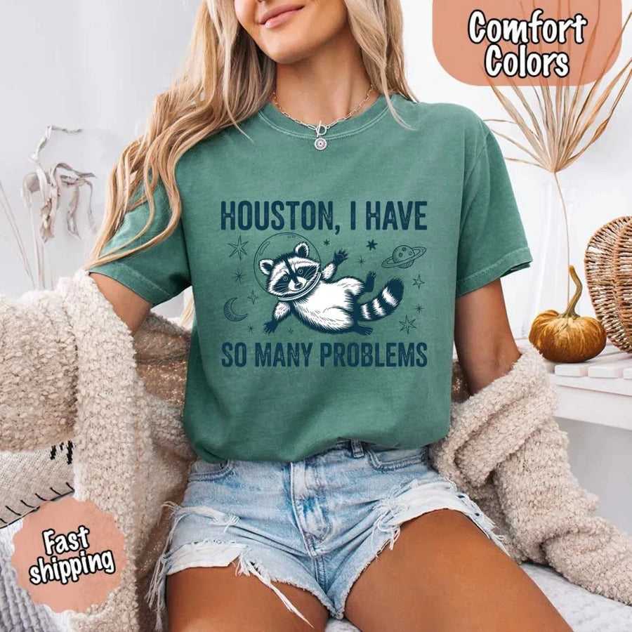 May include a cartoon raccoon in a space helmet floating in space, surrounded by stars, a planet with rings, and a crescent moon. The text reads: “HOUSTON, I HAVE SO MANY PROBLEMS,” parodying the famous Apollo 13 quote with a humorous twist.