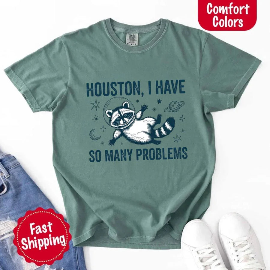 May include a cartoon raccoon in a space helmet floating in space, surrounded by stars, a planet with rings, and a crescent moon. The text reads: “HOUSTON, I HAVE SO MANY PROBLEMS,” parodying the famous Apollo 13 quote with a humorous twist.