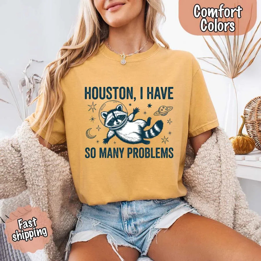 May include a cartoon raccoon in a space helmet floating in space, surrounded by stars, a planet with rings, and a crescent moon. The text reads: “HOUSTON, I HAVE SO MANY PROBLEMS,” parodying the famous Apollo 13 quote with a humorous twist.