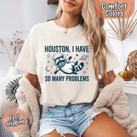 May include a cartoon raccoon in a space helmet floating in space, surrounded by stars, a planet with rings, and a crescent moon. The text reads: “HOUSTON, I HAVE SO MANY PROBLEMS,” parodying the famous Apollo 13 quote with a humorous twist.