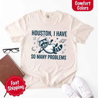 May include a cartoon raccoon in a space helmet floating in space, surrounded by stars, a planet with rings, and a crescent moon. The text reads: “HOUSTON, I HAVE SO MANY PROBLEMS,” parodying the famous Apollo 13 quote with a humorous twist.