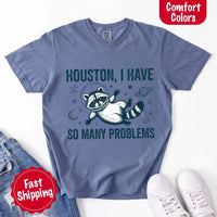 May include a cartoon raccoon in a space helmet floating in space, surrounded by stars, a planet with rings, and a crescent moon. The text reads: “HOUSTON, I HAVE SO MANY PROBLEMS,” parodying the famous Apollo 13 quote with a humorous twist.