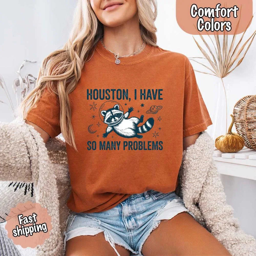 May include a cartoon raccoon in a space helmet floating in space, surrounded by stars, a planet with rings, and a crescent moon. The text reads: “HOUSTON, I HAVE SO MANY PROBLEMS,” parodying the famous Apollo 13 quote with a humorous twist.