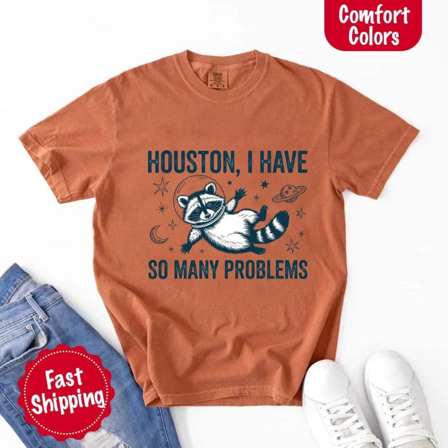 May include a cartoon raccoon in a space helmet floating in space, surrounded by stars, a planet with rings, and a crescent moon. The text reads: “HOUSTON, I HAVE SO MANY PROBLEMS,” parodying the famous Apollo 13 quote with a humorous twist.
