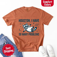 May include a cartoon raccoon in a space helmet floating in space, surrounded by stars, a planet with rings, and a crescent moon. The text reads: “HOUSTON, I HAVE SO MANY PROBLEMS,” parodying the famous Apollo 13 quote with a humorous twist.