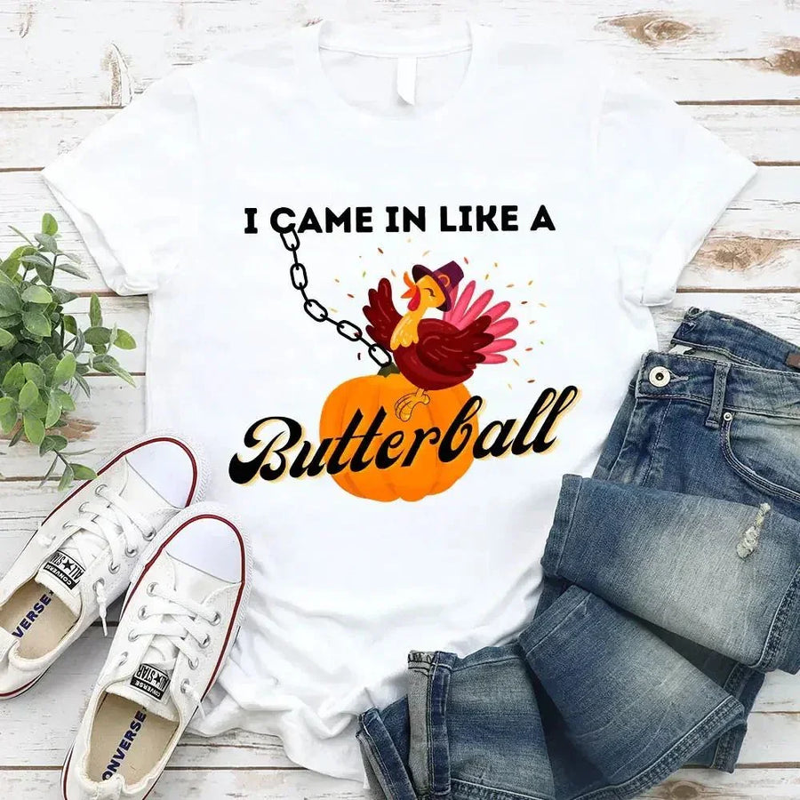 May include a casual crew-neck T-shirt with a playful Thanksgiving graphic showing a turkey stepping onto or perched on a pumpkin, surrounded by leaves or autumn motifs, with bold front text reading “I Came In Like A Butterball.”