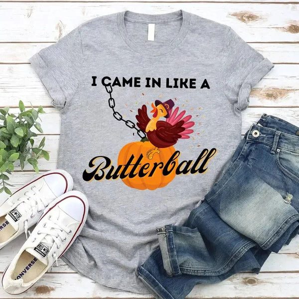 May include a casual crew-neck T-shirt with a playful Thanksgiving graphic showing a turkey stepping onto or perched on a pumpkin, surrounded by leaves or autumn motifs, with bold front text reading “I Came In Like A Butterball.”
