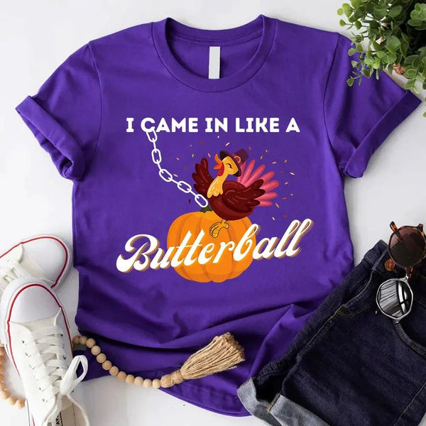 May include a casual crew-neck T-shirt with a playful Thanksgiving graphic showing a turkey stepping onto or perched on a pumpkin, surrounded by leaves or autumn motifs, with bold front text reading “I Came In Like A Butterball.”