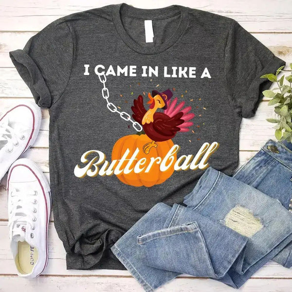 May include a casual crew-neck T-shirt with a playful Thanksgiving graphic showing a turkey stepping onto or perched on a pumpkin, surrounded by leaves or autumn motifs, with bold front text reading “I Came In Like A Butterball.”